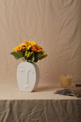 Still life photo of white oval ceramic vase shaped like human face. Modern creative flower vase with sunflowers is located on beige cloth near open book and aromatic candle.  