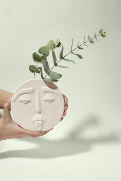 Cropped shot of female hands with white round ceramic vase shaped like human face. Two bluegum twigs are placed in modern creative flower vase.  