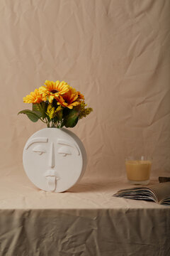 Still life photo of white round ceramic vase shaped like human face. Modern creative flower vase with sunflowers is located on beige cloth near open book and aromatic candle.  