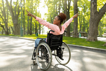 Fototapeta na wymiar Handicapped black woman in wheelchair on walk at city park, showing victory gesture, feeling positive and motivated