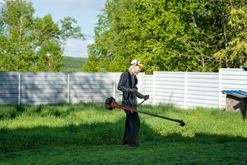 a young man in a protective plastic mask and with a trimmer on his shoulder mowing grass