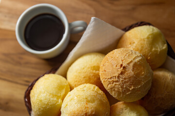 Uma xícara branca cheia de café e uma pequena cesta de vime com pães de queijo sobre uma superfície de madeira.