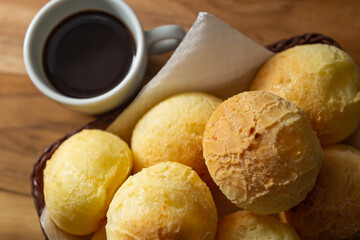 Uma xícara branca cheia de café e uma pequena cesta de vime com pães de queijo sobre uma superfície de madeira.