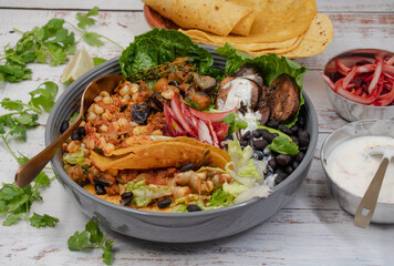 Vegan Mexican taco bowl consisting of spicy corn, black beans, eggplant and mushroom, salad, pickles. White wooden table