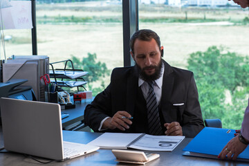 Image of Bearded man wearing shirt tie and black suit sits in office at table, holds pen in his hand. Man with documents and folders working with laptop at desk in office. Lawyer looking document