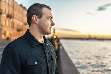 Portrait of a young man watching the sunset on the embankment of the river in the city