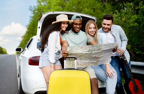Traveling with friends. Happy multiracial young people sitting in open car trunk, checking map, having fun road trip