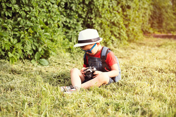 A cute little boy in denim overalls, blue sunglasses and a white hat sits on the grass in the park and holds an old retro camera in his hands.