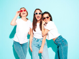 Three young beautiful smiling hipster female in trendy same summer white t-shirt and jeans clothes. Sexy carefree women posing near light blue wall in studio. Cheerful and positive models having fun