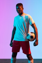 Half-length portrait of African professional football player standing isolated on gradient blue pink background in neon light.