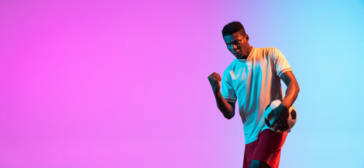 Fototapeta na wymiar African man, professional football player standing isolated on gradient blue pink background in neon light.