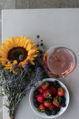 A glass of rose wine stands on the table on the balcony, next to a plate of strawberries and blackberries and a bouquet of flowers made of sunflower and blue thorn. Drink wine. Summer day off. 