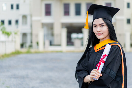 A young beautiful Asian woman university graduate in graduation gown and mortarboard holds a degree certificate stands in front of the university building after participating in college commencement