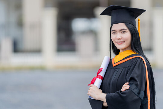 A young beautiful Asian woman university graduate in graduation gown and mortarboard holds a degree certificate stands in front of the university building after participating in college commencement