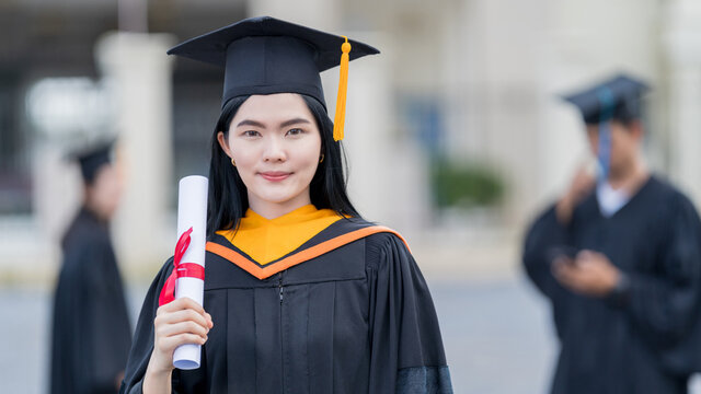 A young beautiful Asian woman university graduate in graduation gown and mortarboard holds a degree certificate stands in front of the university building after participating in college commencement