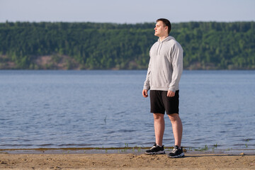 A young man in a gray sweatshirt stretches before jogging on an empty beach at dawn. Slimming and healthy lifestyle concept.