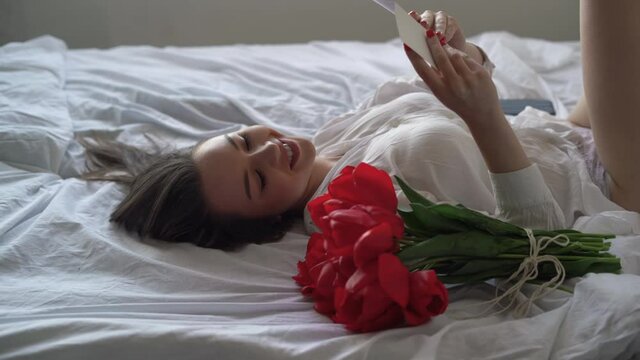 A young Caucasian woman lies in a man's shirt on the bed, red tulips lie nearby. Cheerful smile and reads a note. Holiday gift.