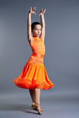 Child girl in an orange sports dress posing in dance movement on gray background