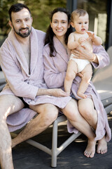 Obraz premium Portrait of a young family with a one year baby boy resting at Spa resort, hug together near the sauna outdoors. Parents with a newborn son on vacation