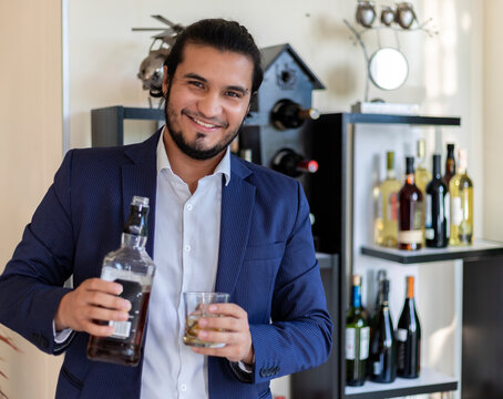 Young Classy Latino Man Wearing Suit, Holding Whiskey Bottle And Glass. Looking At Camera.