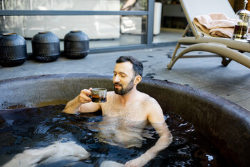 Adult caucasian man relaxing at spa outdoors, lying in a hot vat and drinking tea. Bathing and recreation in Spa