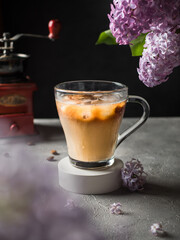 Iced coffee with milk in a glass mug. Lilac flowers, coffee grinder and coffee beans in the background. Dark background