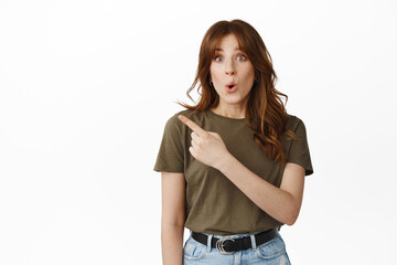 Obraz premium Surprised and excited girl say wow, pointing at upper left corner, showing advertisement with finger, staring in awe at camera, standing against white background