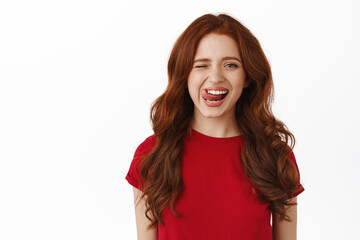 Carefree ginger girl winking, showing tongue and smiling happy, standing in red t-shirt against...