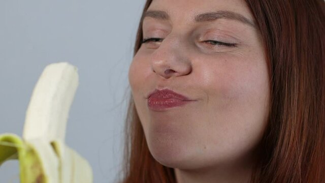 Young woman laughing and eating ripe banana isolated on grey background. Woman biting banana, healthy eating and lifestyle