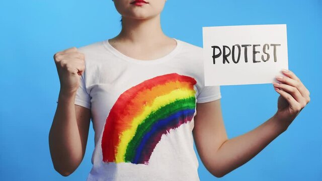 Lgbt protest. Gay discrimination. Anti homophobia transphobia strike. Strong lesbian woman activist with colorful rainbow symbol on t-shirt raising fist placard isolated on blue.