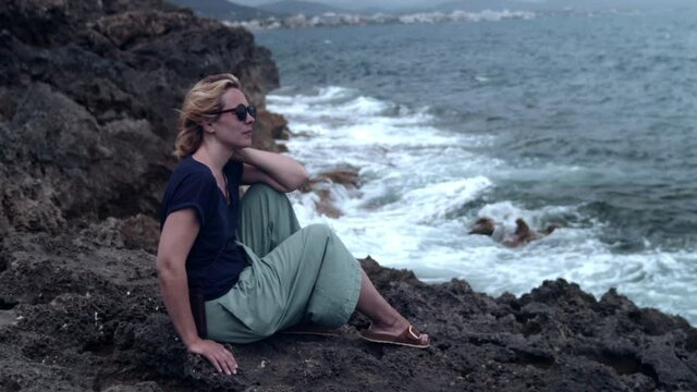 Woman sitting on a cliff and watching the surf of the waves breaking on the rock. Slow motion 60fps_4K