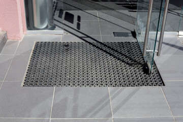 threshold made of gray ceramic tiles at entrance to store with a rubber foot mat and open glass door with metal handle at office building close-up front view, nobody.