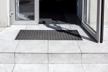 threshold made of light gray ceramic tiles with steps at the entrance to the store with a foot mat and an open glass door at the facade of an office building close-up front view, nobody.