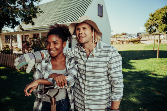 Diverse couple friends smiling into distance showing affection standing in farm village in front of white home dressed in gardening gear 