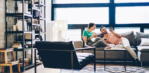 Caucasian hipster guy enjoying weekend leisure for bonding with girlfriend using cellular device for checking status of online booking for together pastime, couple in love communicate in home