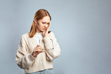 Young tired ill student girl in beige sweater hold glasses put hand on face having headache isolated on gray background. Stressed woman feeling tired after night without sleep.