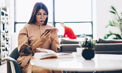 Caucasian hipster girl in trendy eyeglasses using modern smartphone device for making web booking...