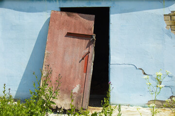 A red, old door with a padlock and a handle covers a dark doorway in the wall. The brick wall is covered with blue plaster. The plaster is falling off the wall. Bricks and cracks on the wall