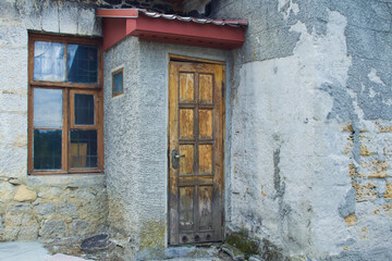 A yellow, wooden, closed door against the background of an  shabby, stone wall. The wall is gray and covered with crumbling plaster. There is an old window in the wall with a grate inside. 