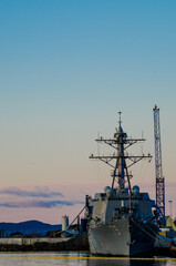 Fototapeta premium An American military ship docked in a foreign harbor with sunset light in the background and industrial buildings