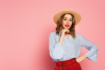 Stylish woman in sun hat holding hand on hip and looking at camera on pink background