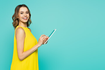 Woman in dress using digital tablet and smiling at camera isolated on blue