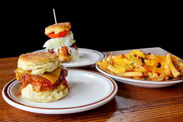 Tasty breakfast / brunch. Biscuit sandwiches (fried chicken, cheese, tomato, blue cheese sauce) and cheesy fries on white plates on the wooden table, black background.