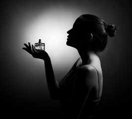 The silhouette of a beautiful young woman in profile with a bottle of perfume in her hand on a dark...