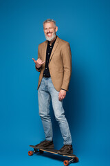 full length of pleased middle aged man riding on longboard and showing call me gesture on blue