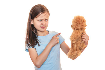 Little girl with a soft toy teddy bear on a white background. The child strictly points her finger...