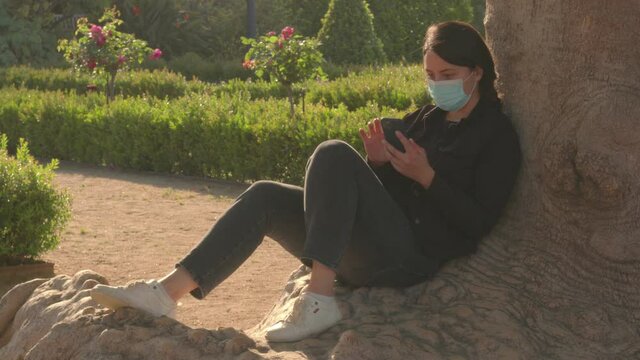 A brunette caucasian girl in black jacket, grey jeans and covid mask is sitting on the tree base in the park enjoy the moment on a sunset. Navigate the internet on the phone. High quality video.