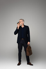 full length of man with grey hair in suit kissing paper cup and holding leather bag on grey
