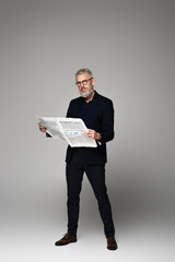 full length of middle aged man in glasses and suit holding economic newspaper on grey