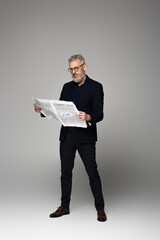 full length of middle aged man in glasses and suit reading economic news on grey
